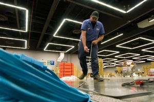 A worker in a blue uniform and cap bends over, handling cables on a patterned metal floor. Bright ceiling lights create a geometric pattern above.