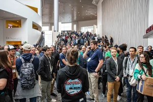 A large crowd gathers in the convention center lobby, many wearing badges. The atmosphere is busy and expectant, with people talking and observing.