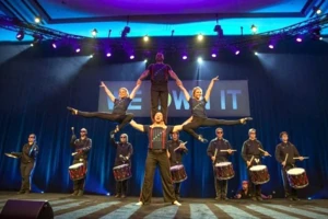 People perform a balancing act with drummers in the background on a stage, under spotlights, in front of a sign that reads "KNOW IT.