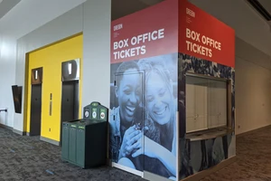 Box office ticket counter with closed window, features a large mural of two people smiling. Bright yellow elevator doors are adjacent on the left.