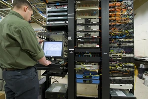 A technician in a green shirt works in a data center, monitoring graphs on a computer screen near large server racks filled with cables and hardware.