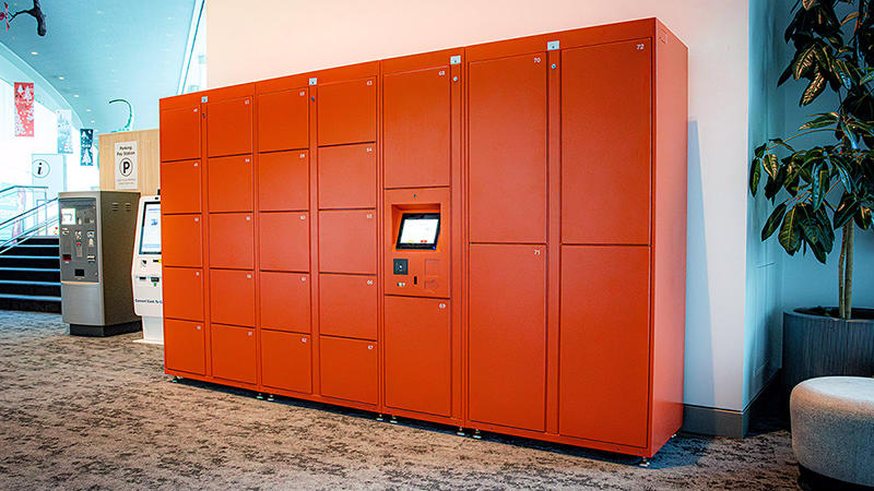 A bank of dark orange lockers in a brightly lit building lobby next to an ATM and parking payment kioski