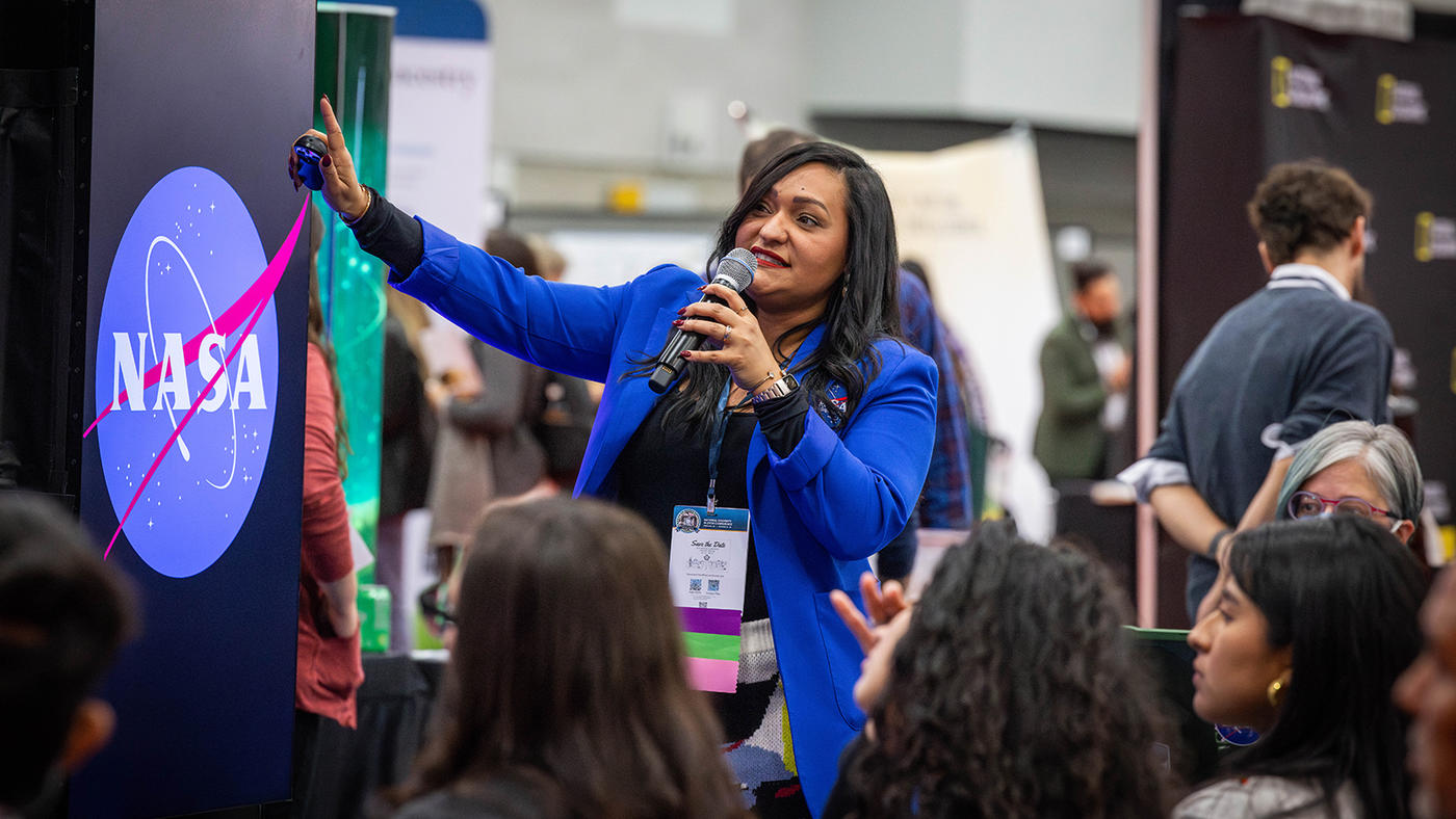 A woman in a blue jacket speaks into a microphone and points at a screen in front of several interested youth