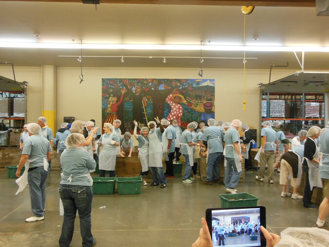 About 35 people in hairnets and aprons during a day of service for the Oregon Food Bank. Two volunteers at the center pose and wave while someone takes their photo.