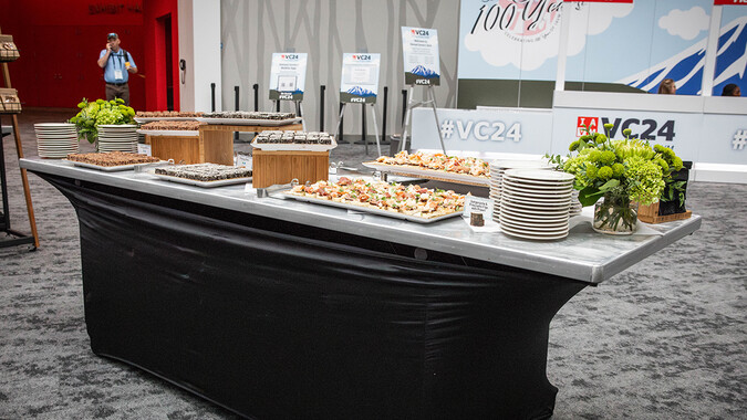 A buffet table with assorted appetizers, stacked plates, and green floral arrangements set up in the convention center.