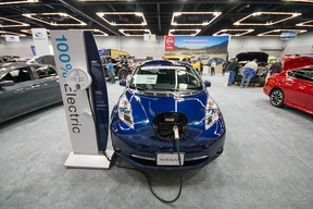 Electric vehicle show scene with a blue electric car charging at a station labeled "100% Electric," surrounded by cars and attendees in a well-lit hall.