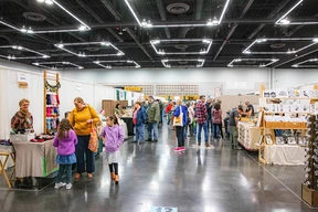 A bustling craft fair with various booths. Visitors, including families, browse colorful handmade items. Bright overhead lighting enhances the lively, engaging atmosphere.