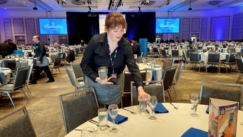 Banquet hall with round tables and blue napkins. A server places a water glass on a table. Room has dim lighting; two screens display "HopeBuilders."
