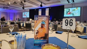 Banquet hall with event tables set for dining. A "Build Stability, Give Today" pamphlet featuring a construction worker is on a table labeled 96. Mood is formal and supportive.